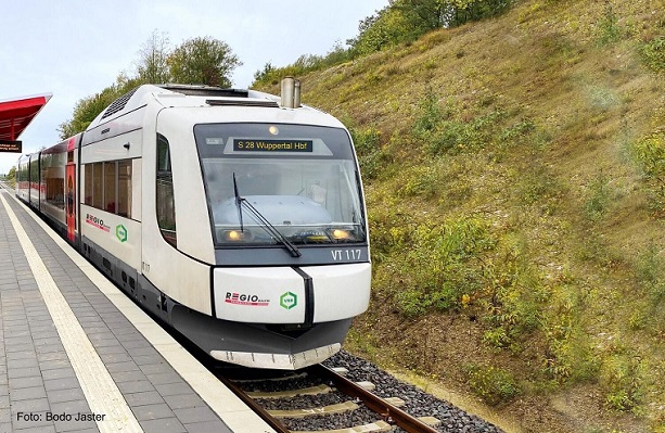 Ein weißer Zug des Typs Integral S5D95 steht an einem Bahnsteig neben einem grünen Wiesenhang.