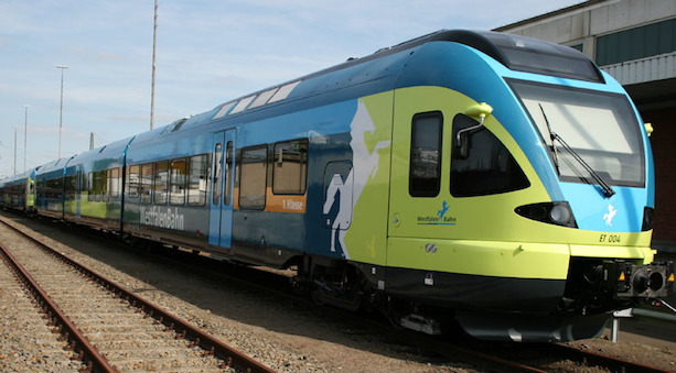 Ein Zug vom Typ Flirt, bunt gestaltet in Blau-, Grün-, Orange- und Weißtönen, steht an einem Bahnsteig.