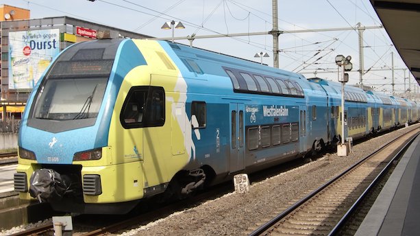 Ein Zug vom Typ KISS, gestaltet in Blau-, Grün- und Weißtönen steht an einem Bahnsteig.