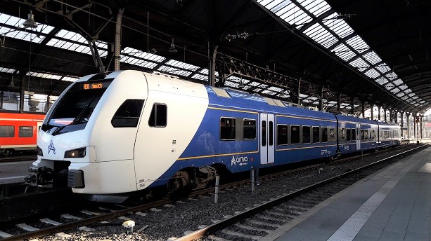 Ein weiß-blauer Zug vom Typ FLIRT3 steht an einem Bahnsteig in einem überdachten Bahnhof.
