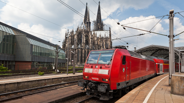 Eine rote Lomotive vom Typ BR 146 zieht rote Doppelstockwagen aus dem Kölner Hauptbahnhof, im Hintergrund der Kölner Dom und das Museum Ludwig.
