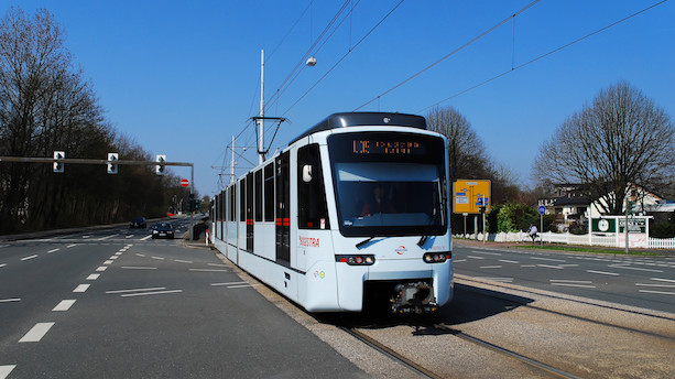 Ein hellblauer Stadtbahnwagen vom Typ Tango fährt über Gleise zwischen zwei Straßen.