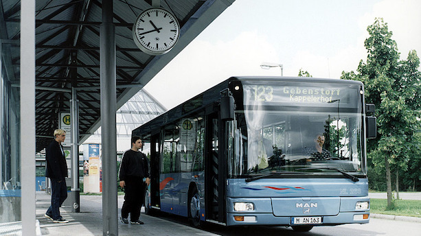 Ein Standardlinienbus für den Stadtverkehr steht mit geöffneten Türen an einem Busbahnhof, zwei Jugendliche sind im Begriff, einzusteigen.