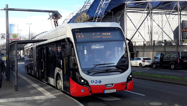 Ein weiß-roter Batterielinienbus steht an einer Haltestelle und ist an eine Batterieladevorrichtung angeschlossen.