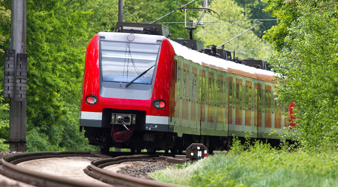 Ein rot-weißer Zug fährt über Gleise, die auf beiden Seiten von dichter Begrünung umgeben sind.