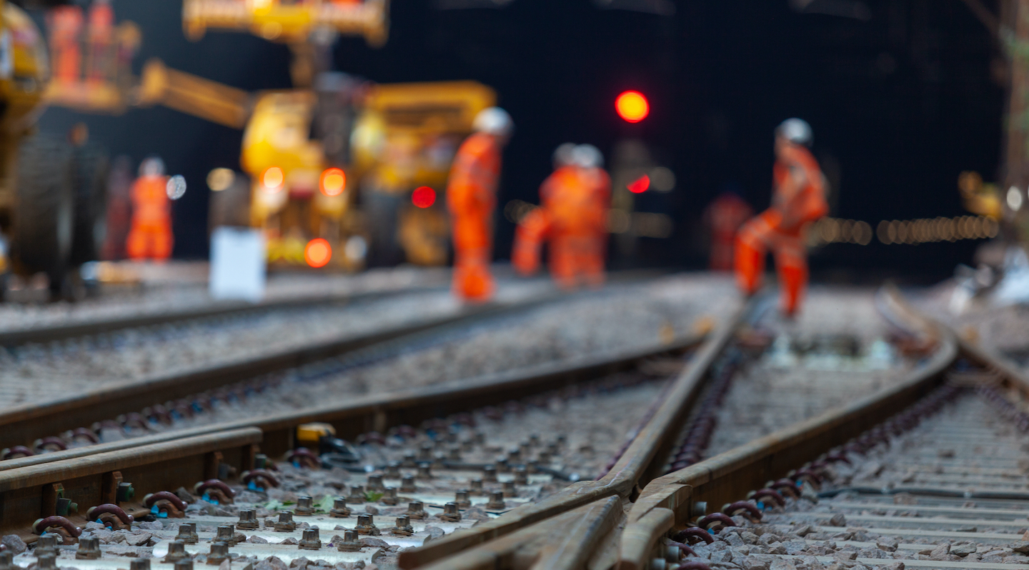 Im Vordergrund Bahnschienen, sehr unscharf im Hintergrund zu erkennen Bauarbeiten vor einem Tunnel mit orange gekleideten Arbeitenden und großen gelben Maschinen.