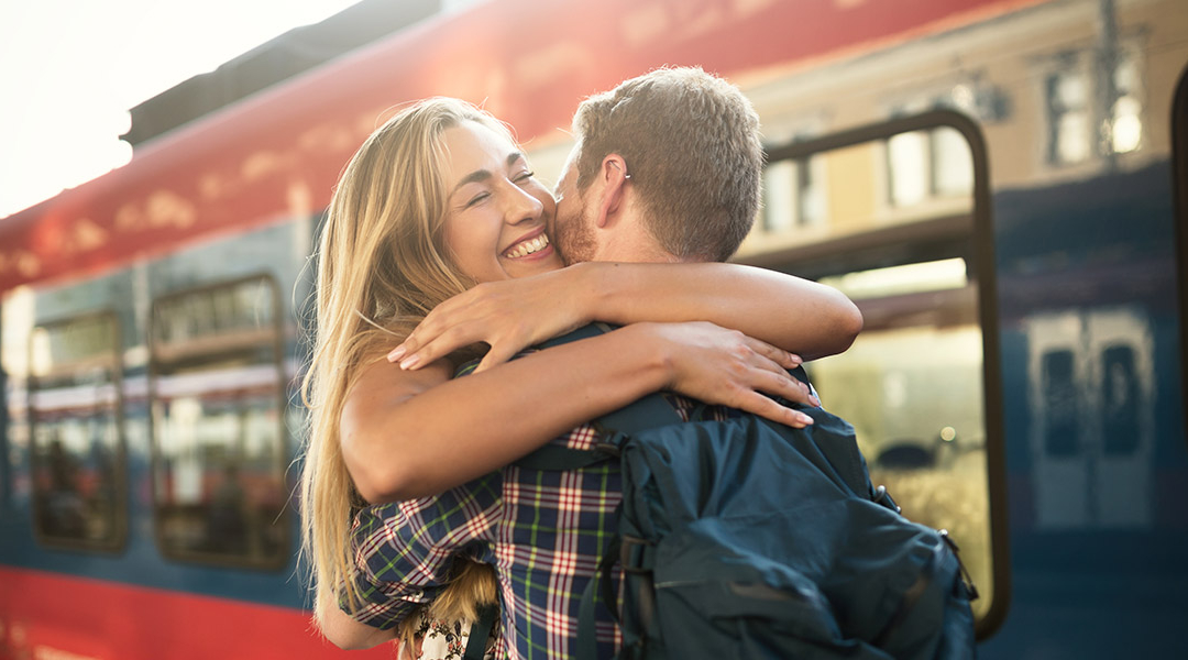 Ein Mann und eine Frau umarmen sich innig und wirken glücklich, im Hintergrund ein Zug.
