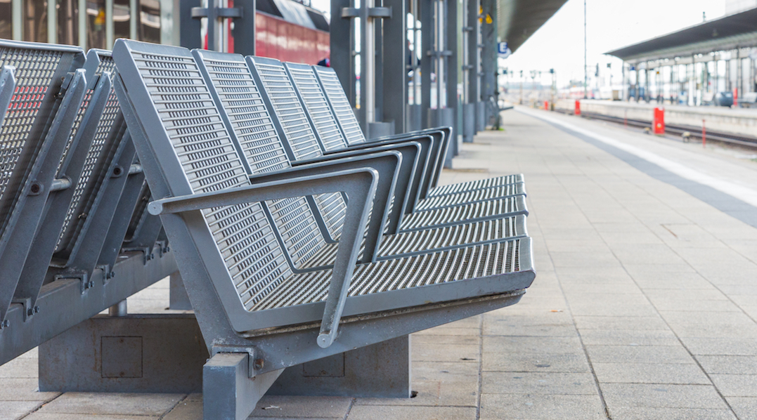 Der Blick über die Länge eines leeren Bahnsteiges, im Vordergrund eine grau lackierte Metall-Bank mit fünf Sitzgelegenheiten, die mit Armlehnen abgetrennt sind