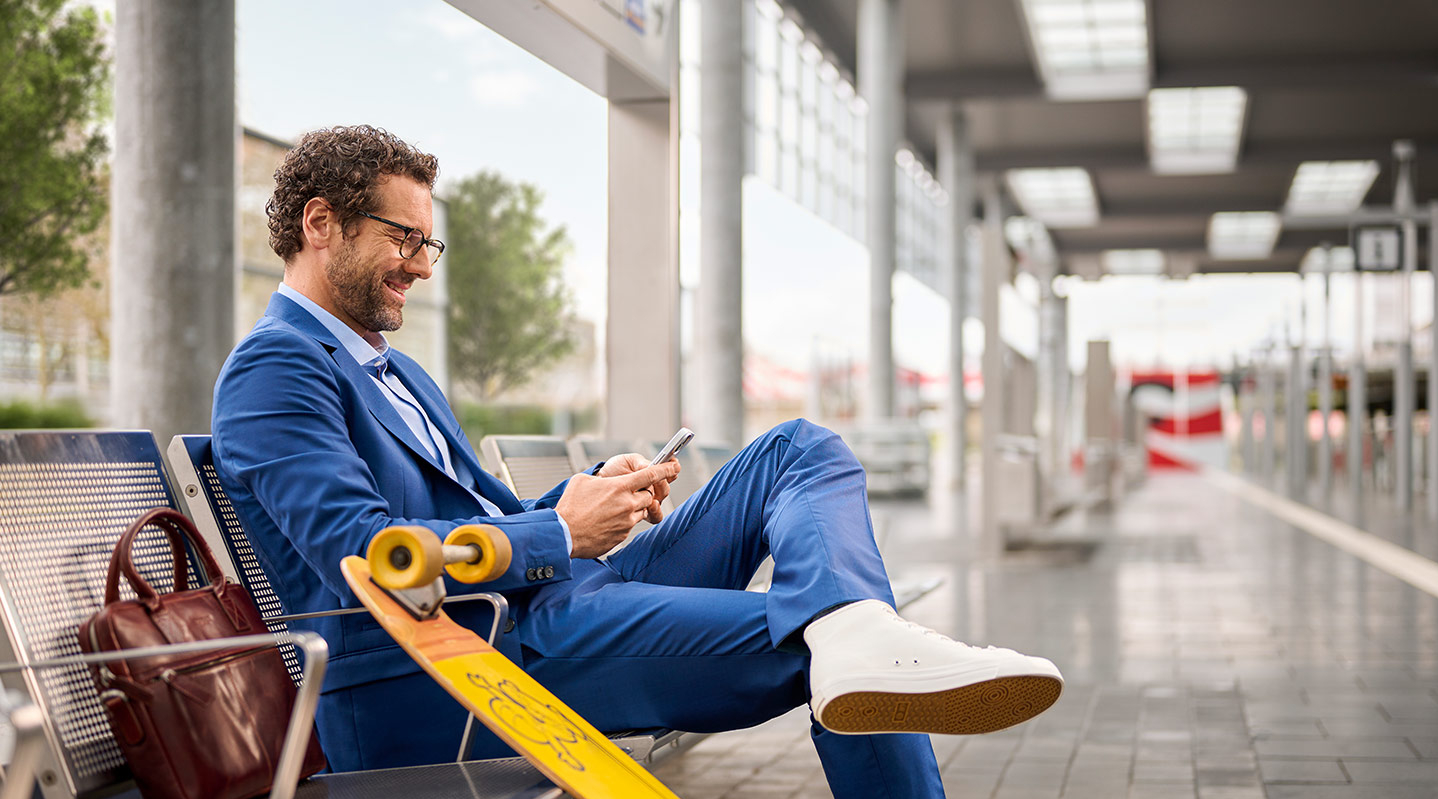 Ein sportlicher Business-Mann sitzt auf einer Bank am Bahnsteig und schaut lächelnd auf sein Handy.