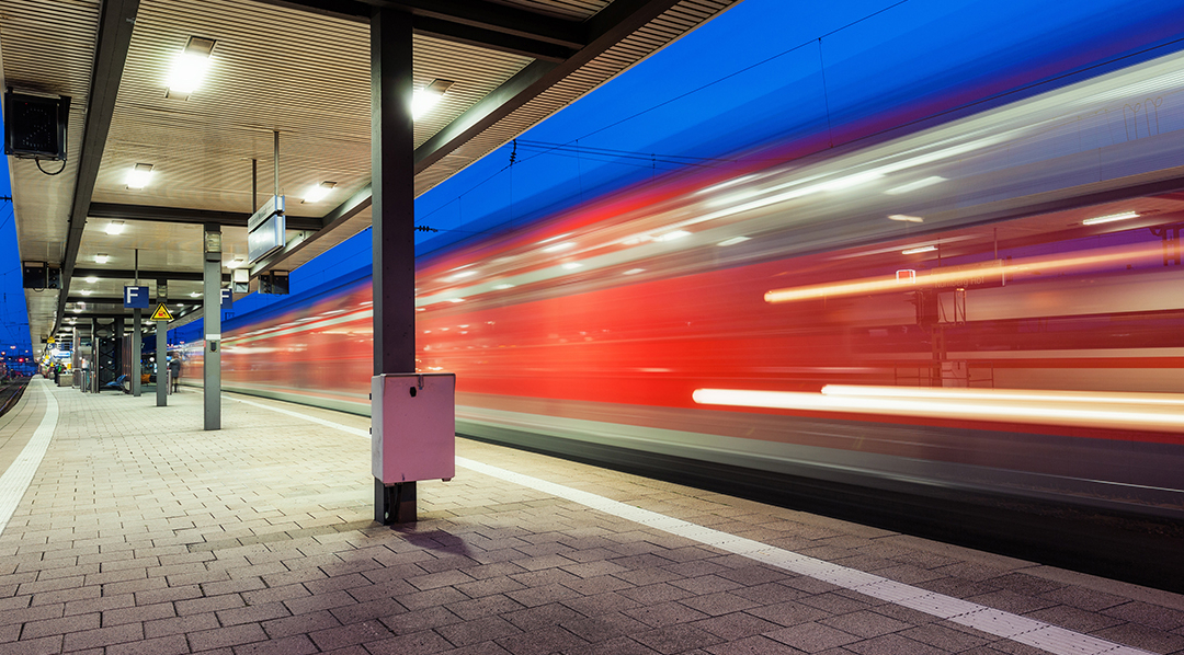 Der Blick über einen Bahnsteig am Abend, während an einem Gleis ein hell beleuchteter Zug mit Bewegungsunschärfe abfährt