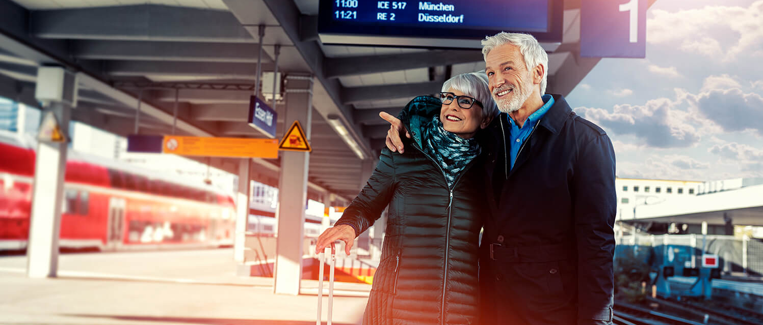 Ein älteres Ehepaar steht mit Koffern an einem bahnsteig und blickt die Gleise entlang.