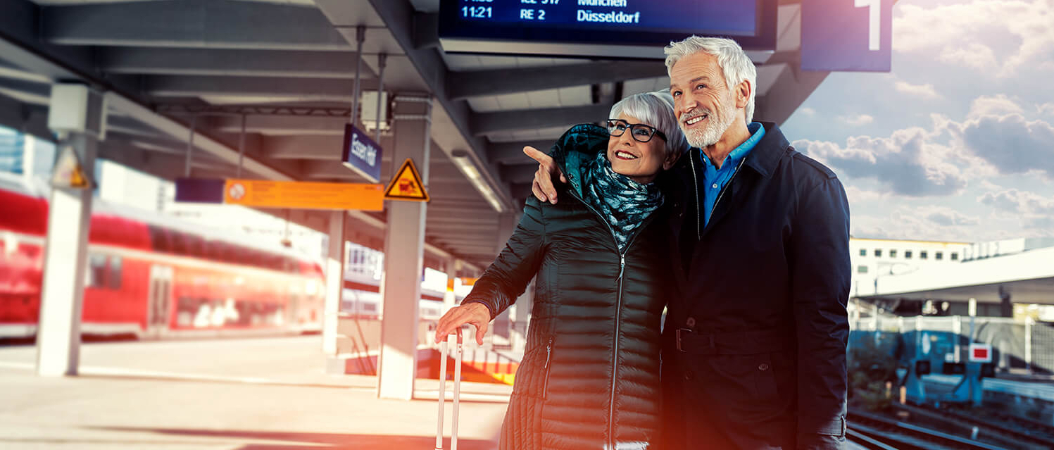 Ein älteres Ehepaar steht mit Koffern an einem bahnsteig und blickt die Gleise entlang.