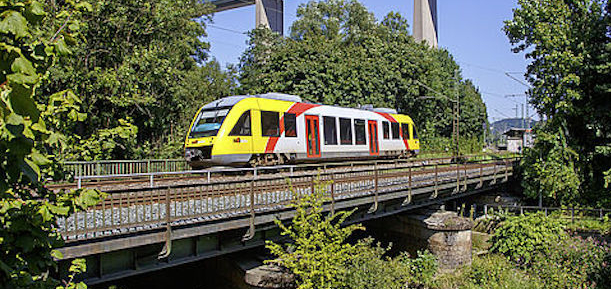 Ein gelb-rot-weiß gestalteter Dieseltriebwagenzug des Typs BR640 bei der Fahrt über eine Flussbrücke in einer grünen Landschaft.