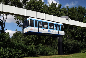 Ein Fahrzeug der H-Bahn hängt an einem unten geschlitzten Hohlkastenträger, in dessen Innern Antrieb, Führung und Stromversorgung wettergeschützt untergebracht sind.