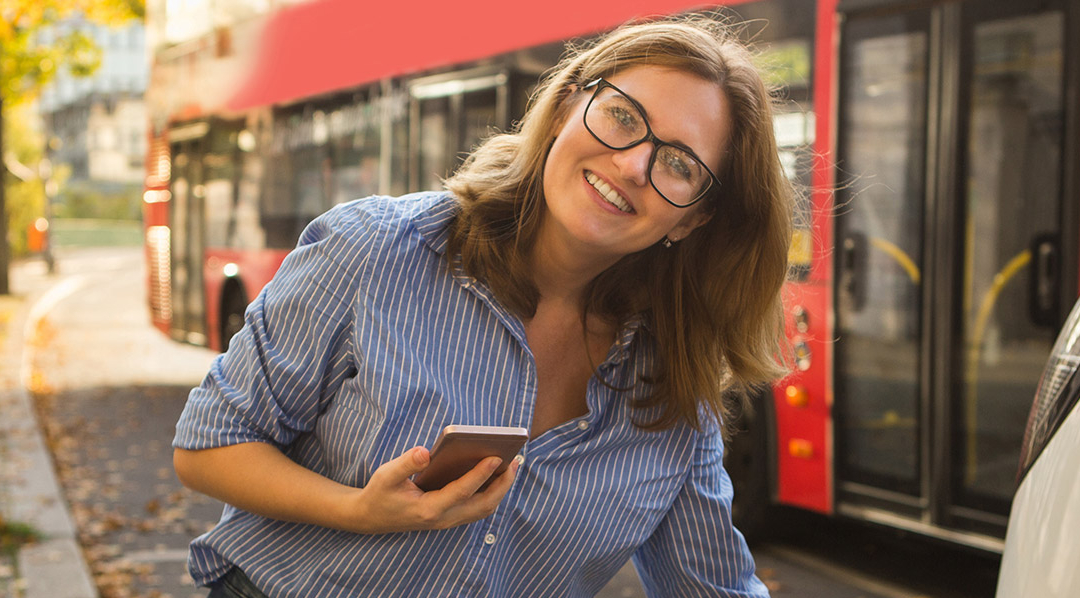 Eine Frau mit Brille lächelt in die Kamera, während sie an einer Ladestation ein E-Auto lädt und hält dabei ihr Smartphone in der Hand. Hinter ihr fährt ein Bus los.