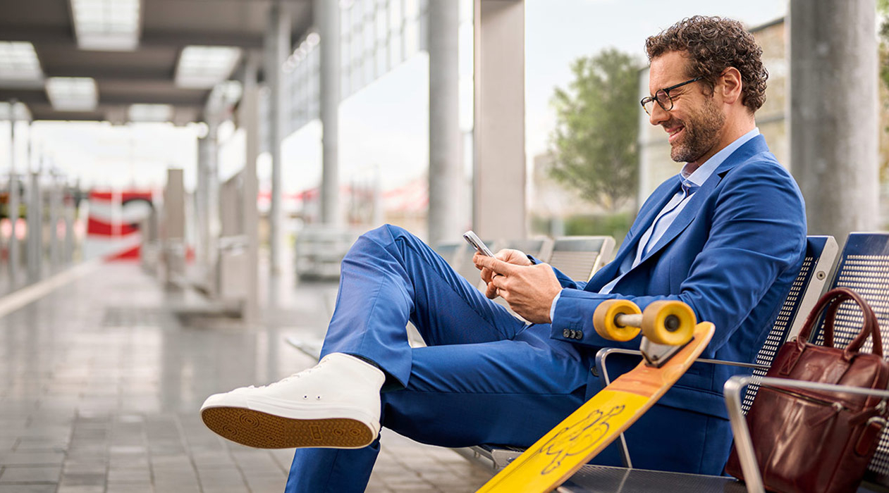 Ein Businessmann sitzt an einem Bahnsteig und schaut lächelnd auf sein Handy.