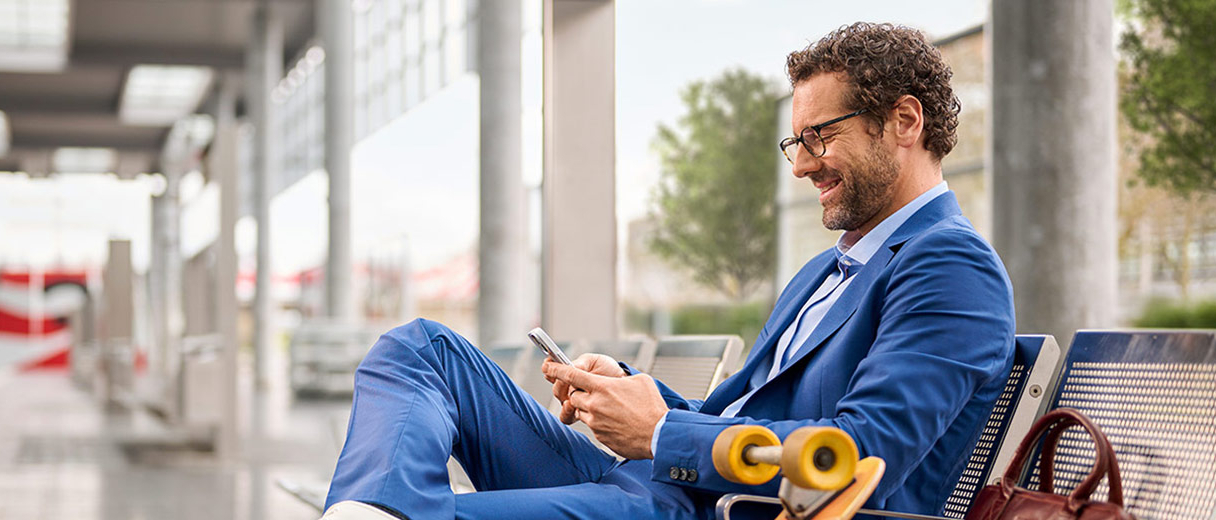 Ein Businessmann sitzt an einem Bahnsteig und schaut lächelnd auf sein Handy.