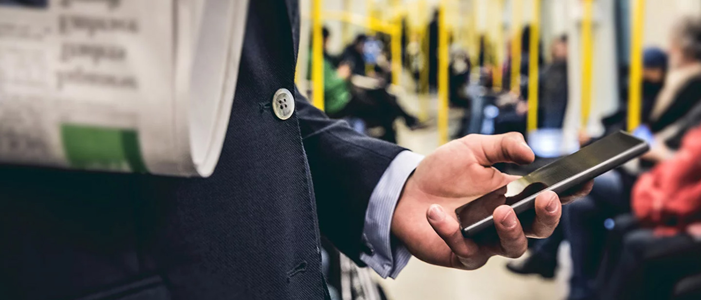 Eine Hand, die ein Smarphone hält, im Vordergrund wird eine Zeitung gehalten, im Hintergrund sieht man den Innenraum samt Passagiere einer Straßenbahn.