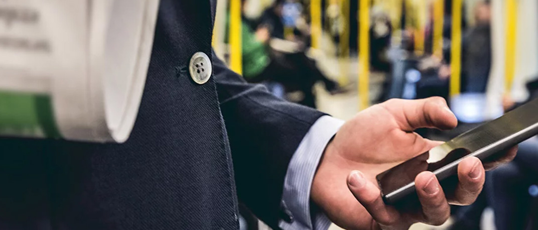 Eine Hand, die ein Smarphone hält, im Vordergrund wird eine Zeitung gehalten, im Hintergrund sieht man den Innenraum samt Passagiere einer Straßenbahn.