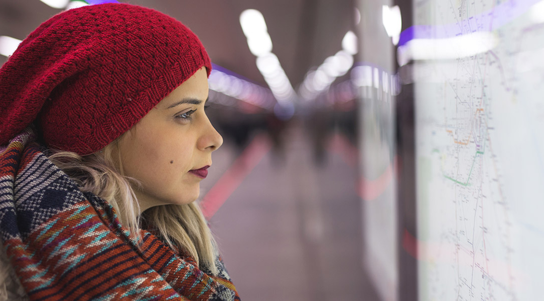 Junge Frau mit Strickmütze und Schal in einem Bahnhof sieht auf ein ausgehängtes Liniennetz