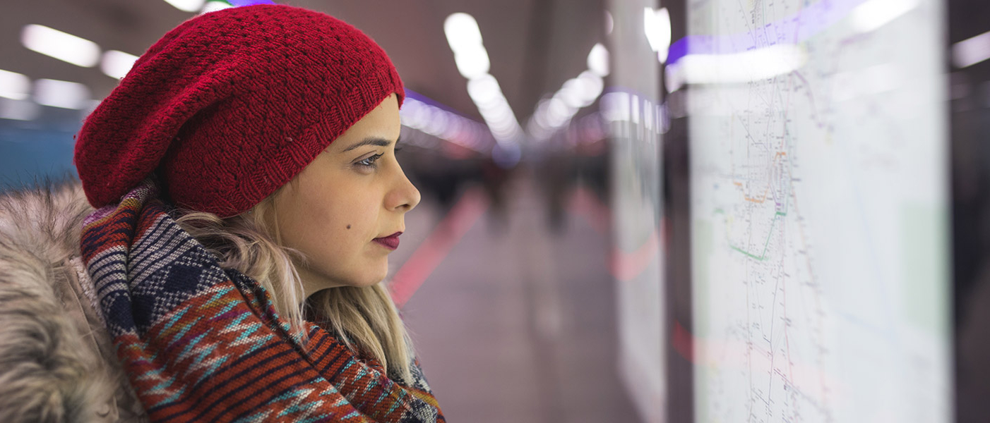 Junge Frau mit Strickmütze und Schal in einem Bahnhof sieht auf ein ausgehängtes Liniennetz