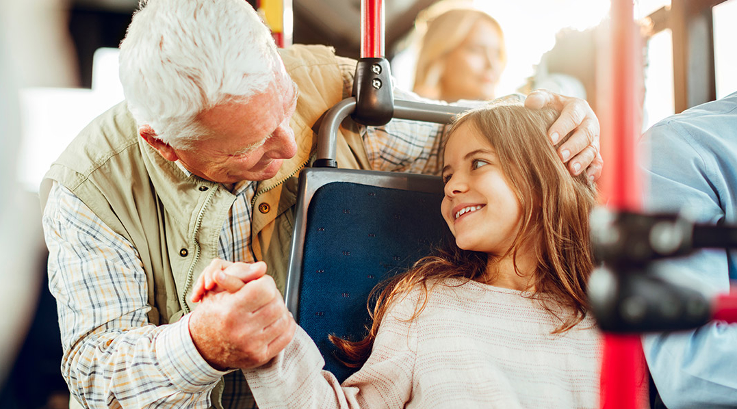 Ein älterer Mann mit weißem Haar sitzt in einem Bus hinter einem jungen Mädchen, beide lächeln sich freudig an, halten sich an den Händen, der Mann hält den Kopf des Mädchens.