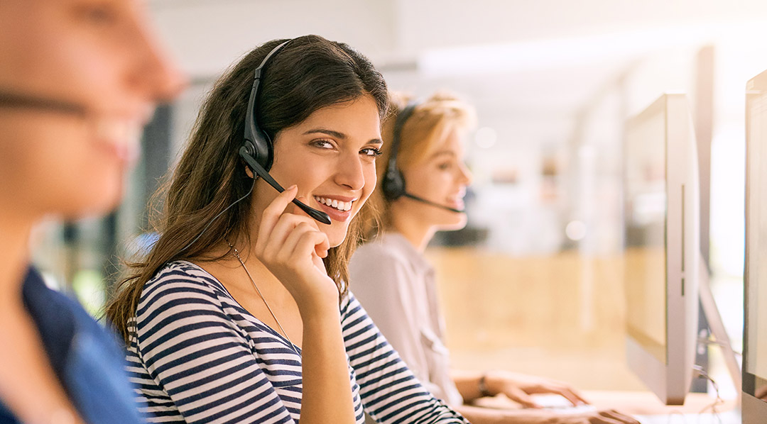 Drei Mitarbeiterinnen eines Callcenters mit Headsets vor Computern, wobei nur die mittig sitzende Frau im Fokus steht und mit direktem Blick in die Kamera lächelt.