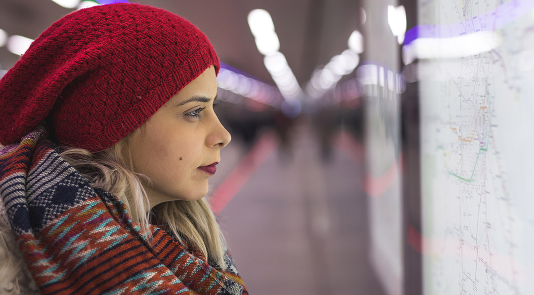 Junge Frau mit Strickmütze und Schal in einem Bahnhof sieht auf ein ausgehängtes Liniennetz