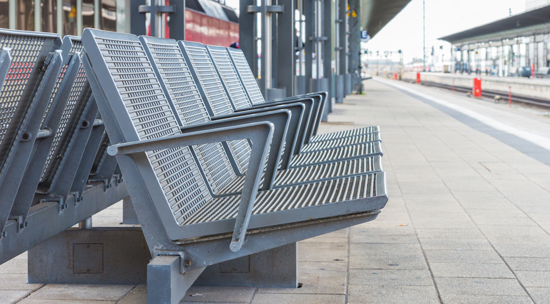 Der Blick über die Länge eines leeren Bahnsteiges, im Vordergrund eine grau lackierte Metall-Bank mit fünf Sitzgelegenheiten, die mit Armlehnen abgetrennt sind