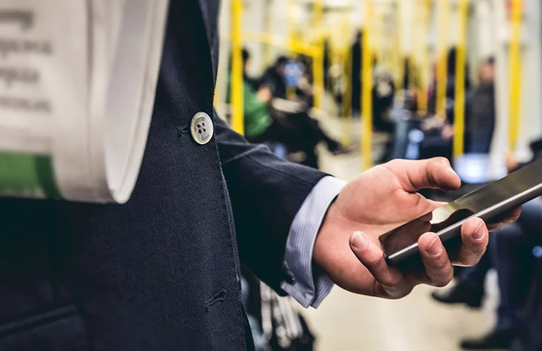 Eine Hand, die ein Smarphone hält, im Vordergrund wird eine Zeitung gehalten, im Hintergrund sieht man den Innenraum samt Passagiere einer Straßenbahn.