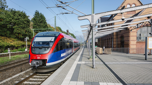 Ein rot-blau gestalteter Dieseltriebwagenug vom Typ BR643.2 steht an einem Bahnsteig eines kleinen Bahnhofs.