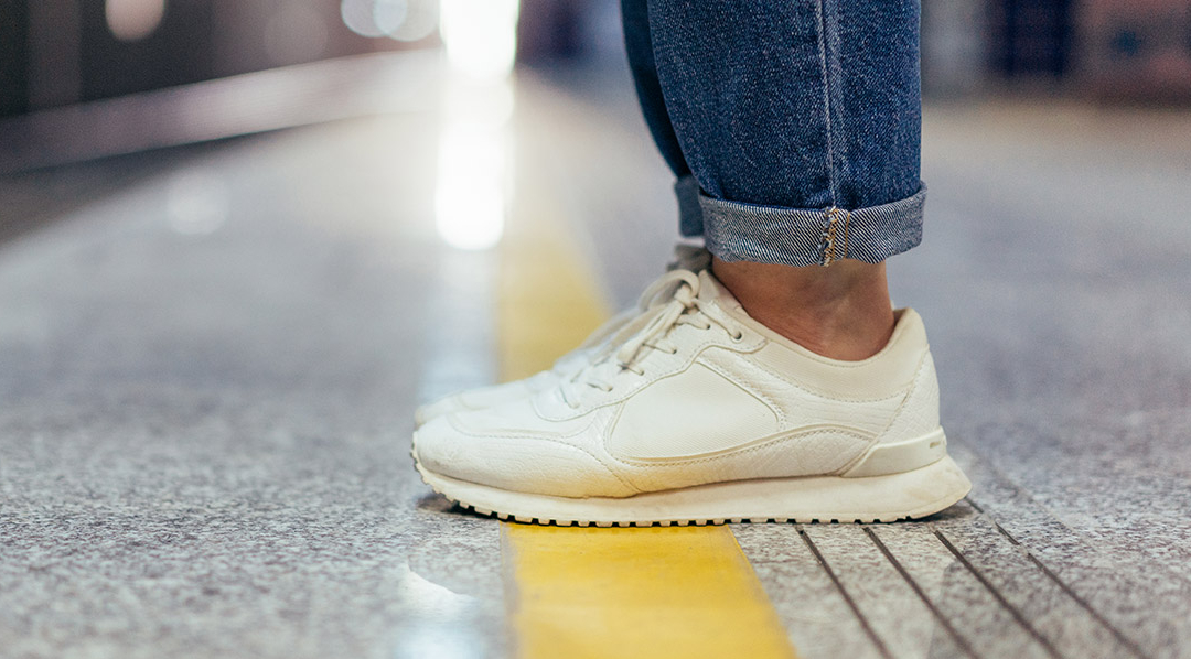 Eine Person steht auf der gelben Linie an einem U-Bahnsteig, sichtbar sind nur die weißen Turnschuhe und etwas von der hochgekrempelten blaue Jeans.