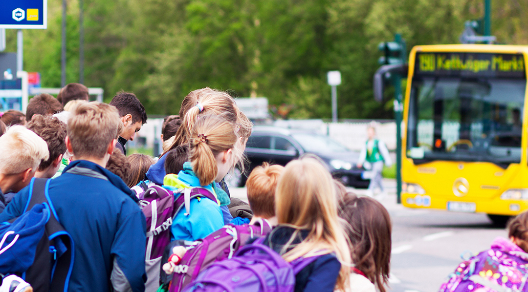 Eine Gruppe Schüler*innen wartet dicht gedrängt an einer Bushaltestelle, während ein gelber Bus in die Richtung der Bushaltestelle einbiegt